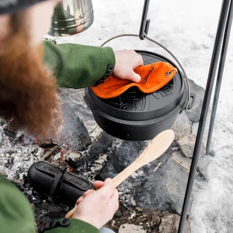 Person handling Petromax cast iron Dutch oven over campfire for outdoor cooking