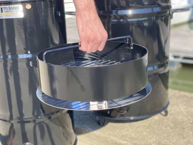 Pit Barrel Ash Catcher attached to charcoal basket, shown being lifted outdoors for convenient ash disposal after grilling.