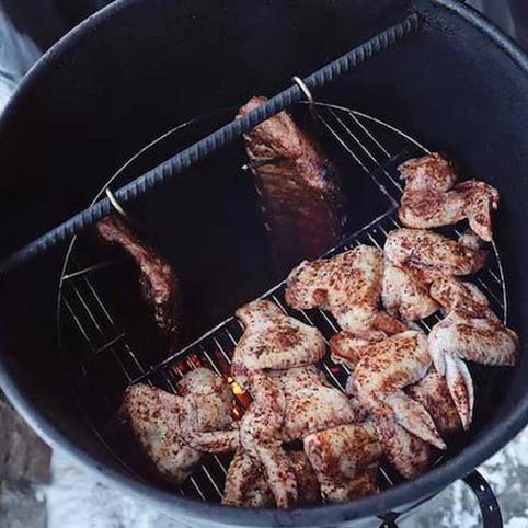 Pit Barrel Hinged Grate loaded with seasoned chicken wings and hanging ribs, demonstrating dual cooking zones in a vertical smoker.