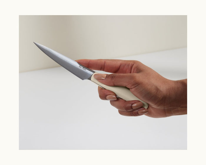 A hand holds the Precise Paring Knife, featuring a white handle and an ultra-sharp German steel blade, set against a neutral background.
