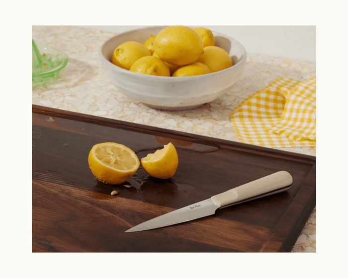 A halved lemon and the Precise Paring Knife rest on a wooden cutting board, with a bowl of whole lemons and a yellow checkered cloth in the background.