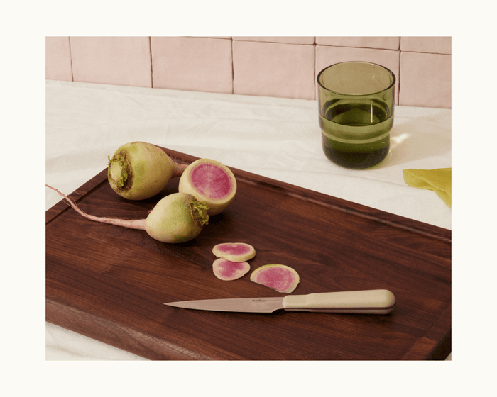 A wooden cutting board with the Precise Paring Knife, two whole watermelon radishes, sliced radishes, and a green glass sits on a white tablecloth.