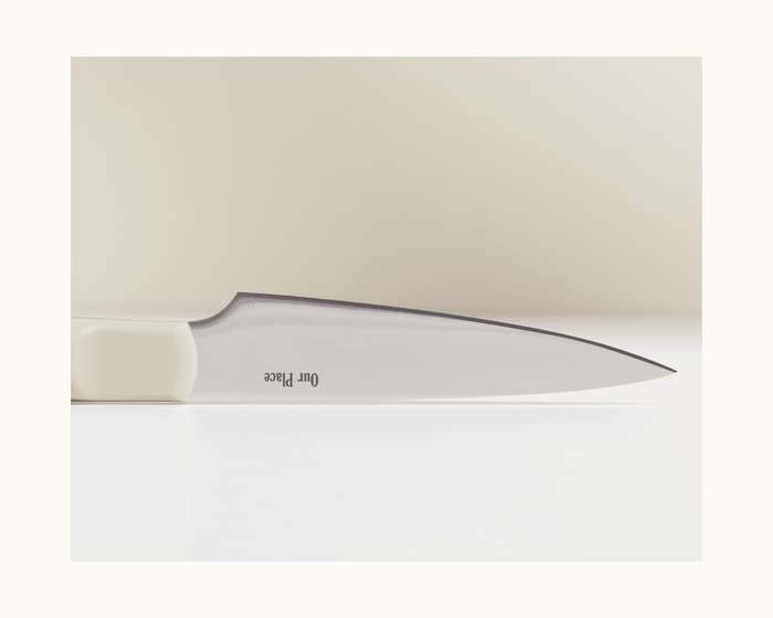 A close-up of the Precise Paring Knife shows its ultra-sharp German steel blade with "Our Place" engraved near the white handle, lying horizontally on a light surface.