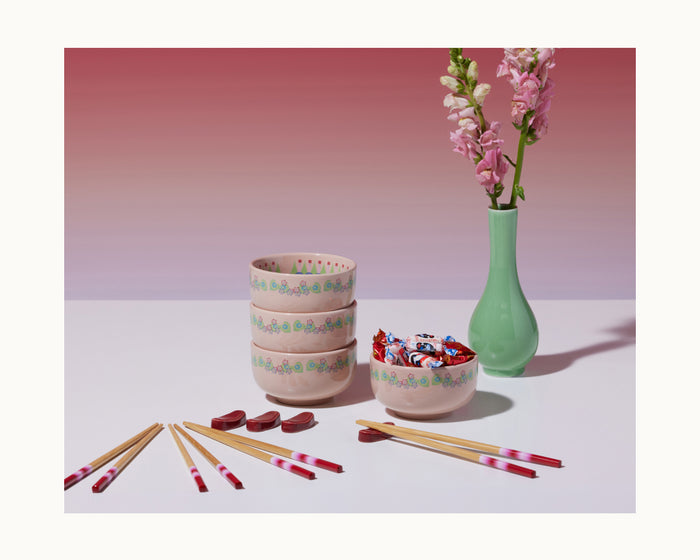 The limited-edition Rice Bowl Set features pink bowls, chopsticks, and heart-shaped rests beside a green vase with pink flowers on a white surface against a pink gradient background.