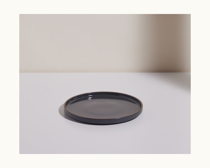 A simple, round, dark gray ceramic Salad Plate sits on a white surface against a neutral background, highlighting the elegance of hand-glazed modern tableware.