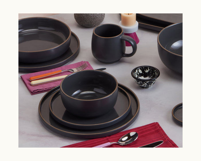 A table set with dark ceramic Salad Plates, bowls, and mugs on pink and red napkins, along with cutlery and a small black-and-white patterned bowl.