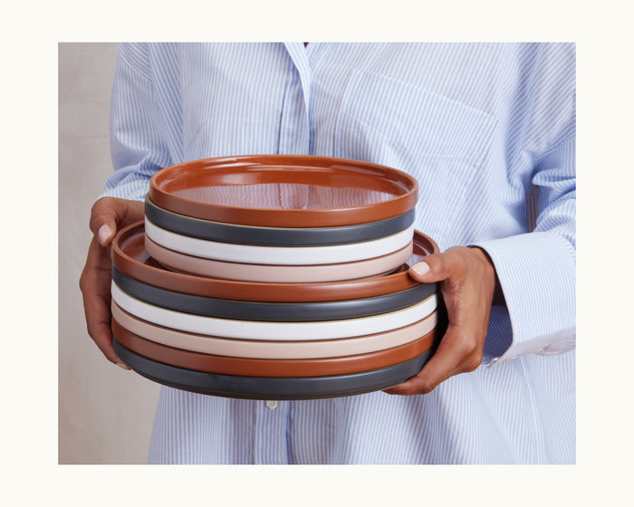 A person in a blue striped shirt holds a stack of Salad Plates—hand-glazed ceramic tableware in brown, black, and white, perfect for any setting.