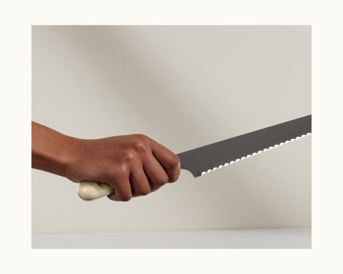 A hand holds the Serrated Slicing Knife with a white handle, made from premium German steel, featuring an ultra-sharp edge, against a plain background.
