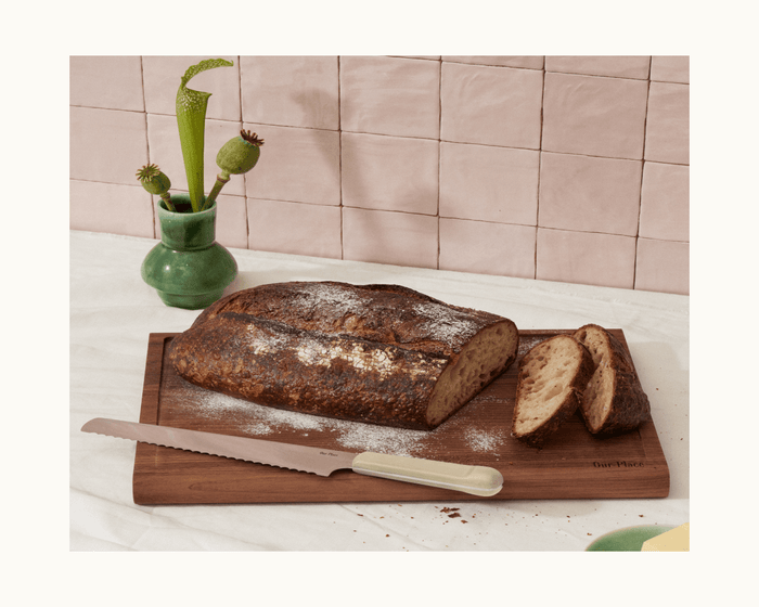 A loaf of rustic bread and two slices rest on a wooden cutting board with a Serrated Slicing Knife beside them; a green vase with plants is in the background.