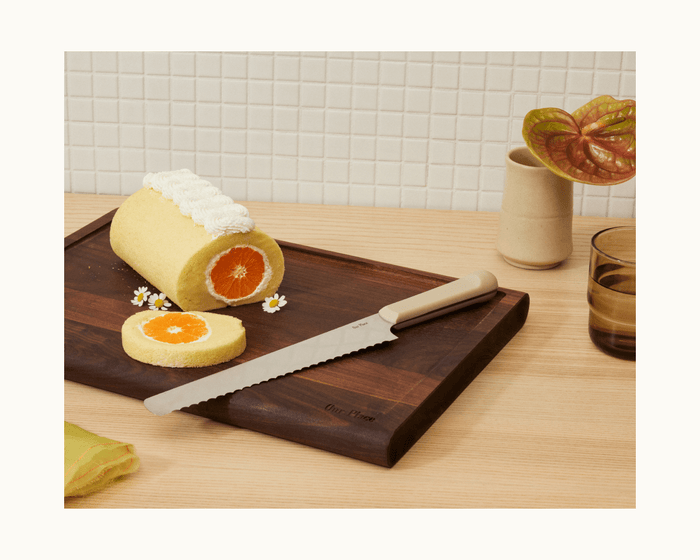 A sliced orange roll cake on a wooden board with a Serrated Slicing Knife, a small vase with a plant, and a glass on a light wood table.