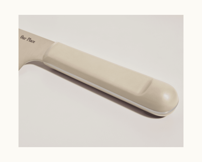 Close-up of the Serrated Slicing Knife's beige handle with "Our Place" engraved on its premium German steel blade, photographed on a light background.