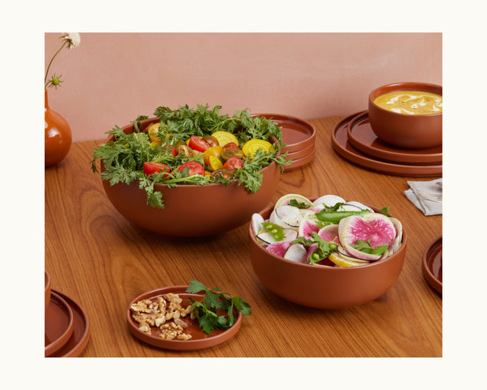 A wooden table set with Serving Bowls filled with salad, sliced vegetables, walnuts, soup, and a small vase holding a single white flower.