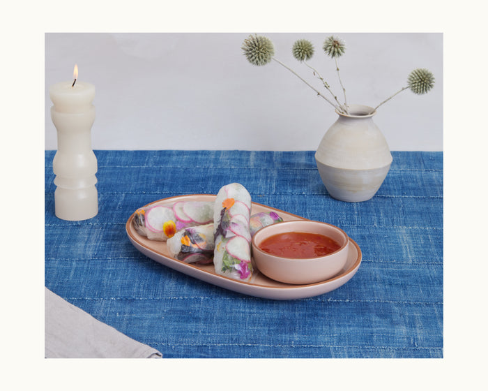 A Serving Platters display features two fresh spring rolls with dipping sauce on a blue tablecloth, accompanied by a lit candle and a ceramic vase with dried flowers for an elegant presentation.