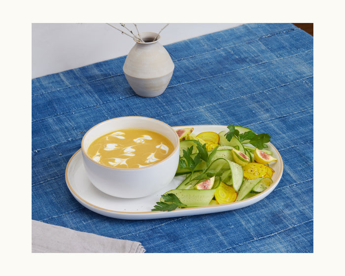 A bowl of yellow soup with white swirls rests on a serving platter, next to a fresh salad of cucumbers, figs, tomatoes, and herbs. A ceramic vase and curved Serving Platters complete the table setting on a blue tablecloth.