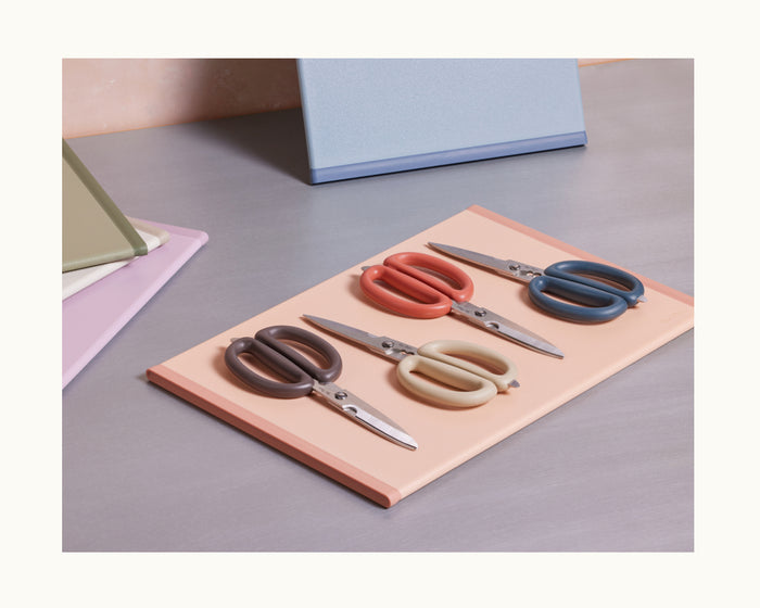 Four pairs of Shear Genius scissors with brown, red, cream, and blue handles, featuring a multifunctional design, are arranged on a closed peach-colored notebook on a desk with other notebooks in the background.