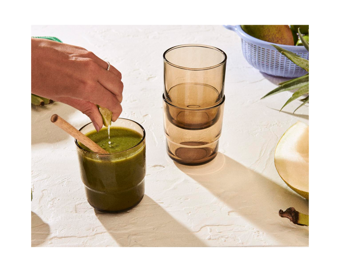 A hand stirs a green smoothie in a glass with a wooden spoon; three stacked Short Night + Day Glasses and fruit are nearby on the table.