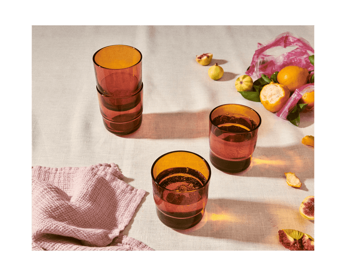 Three Short Night + Day Glasses in amber rest on a light tablecloth, with a pink cloth, assorted fruits, and a pink mesh bag in the background.