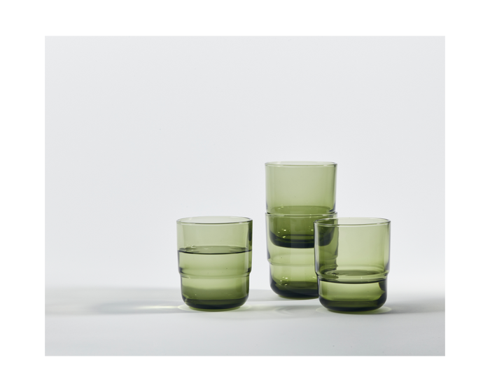 Three Short Night + Day Glasses with a green tint, two half-filled with water, are set on a plain white surface, casting soft shadows.