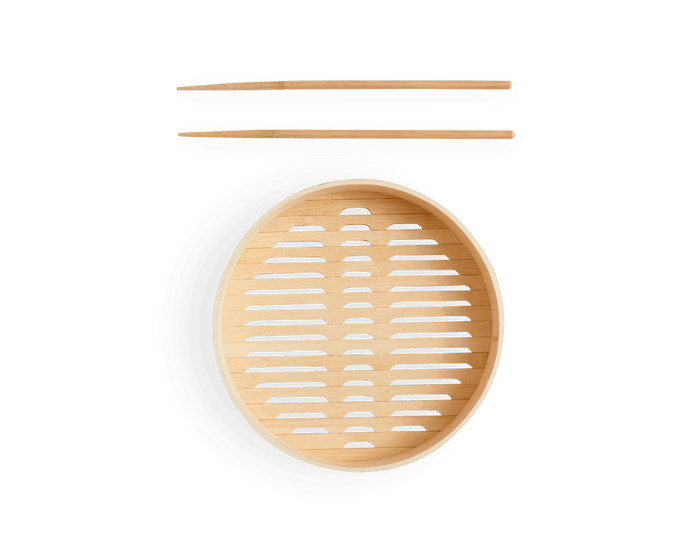 The Spruce Steamer, a round bamboo steamer basket with a slatted base, is placed below bamboo cooking chopsticks against a white background.