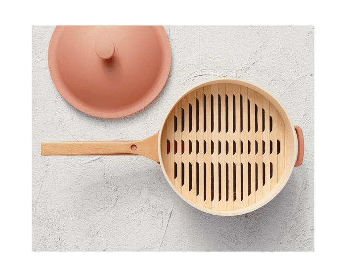 A Spruce Steamer with a wooden handle and slotted steamer insert sits alongside its matching beige and pink lid on a light textured surface.