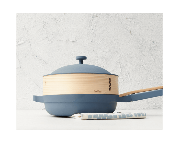 The Spruce Steamer, paired with a blue Always Pan, matching lid, and wooden handle, rests on a white surface with a striped cloth beneath it against a textured white background.