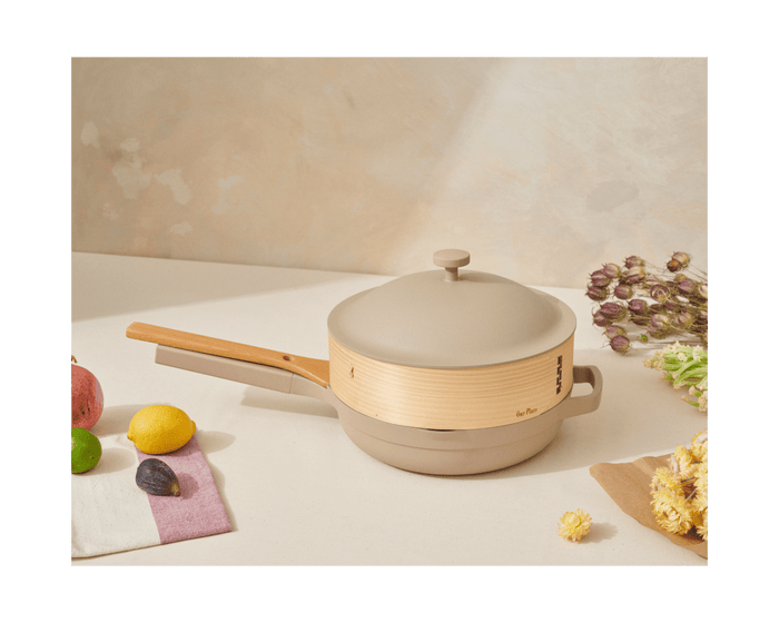 The Spruce Steamer, in beige and light wood with a lid, sits on a countertop surrounded by assorted fruits, flowers, bamboo cooking chopsticks, and a white napkin with purple trim.