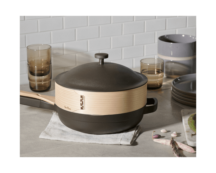 The Spruce Steamer, a lidded cast iron pan with a wooden handle, rests on a folded cloth and is surrounded by glasses, bowls, plates, and bamboo chopsticks on a kitchen counter with a tiled backsplash.