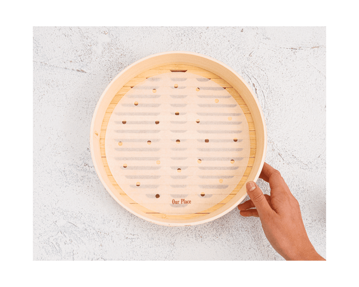 A hand holds the Spruce Steamer, a round basket lined with parchment paper, resting on a light textured surface.