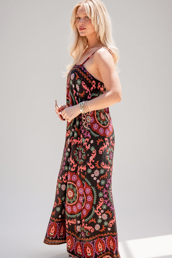 A woman modeling a sleeveless maxi dress with a black base and a vibrant, multicolored bohemian paisley and floral pattern, set against a plain light gray studio background.