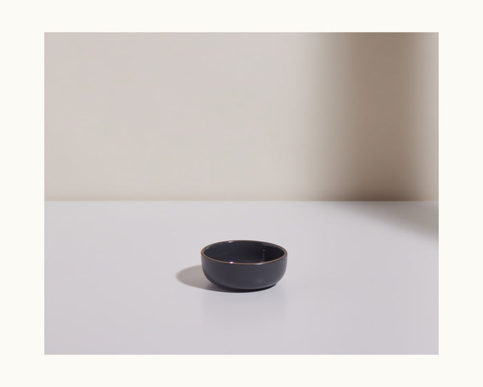 A Tiny Bowls ceramic bowl, small and round with a dark finish, from a modern tableware set is displayed on a white surface against a plain light background.