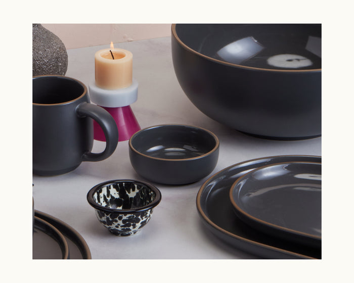 Tiny Bowls, a set of dark ceramic and stackable bowls, is paired with a mug and a black-and-white patterned cup for an elegant tableware display, complemented by a candle and decorative accents on the table.