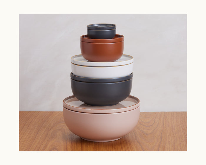 A stack of five colorful Tiny Bowls in assorted sizes rests on a wooden surface with a neutral background, highlighting this versatile ceramic tableware set.