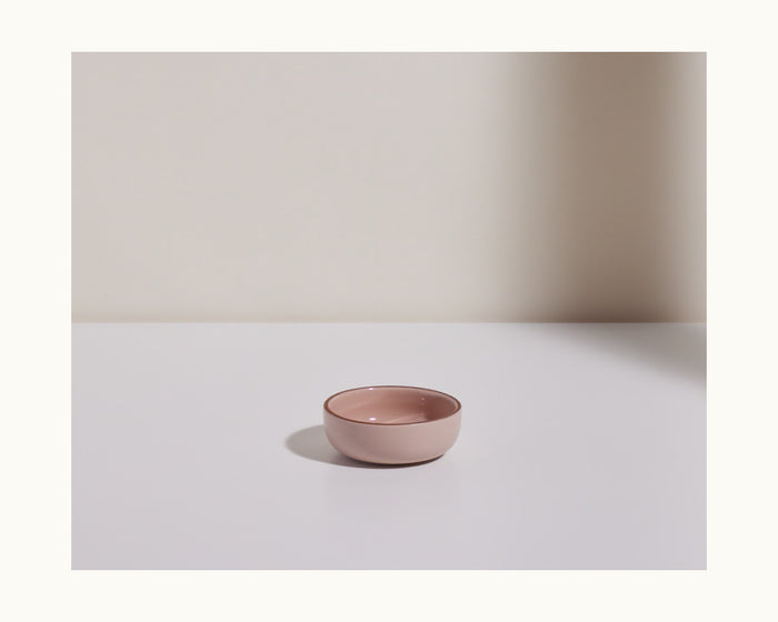 A Tiny Bowls ceramic piece, small, round, and pale pink, from a stylish tableware set rests on a white surface with a neutral background and soft lighting.