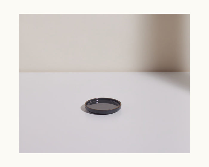 A Tiny Plates small, round black ceramic plate rests on a white surface, casting a shadow to the right.
