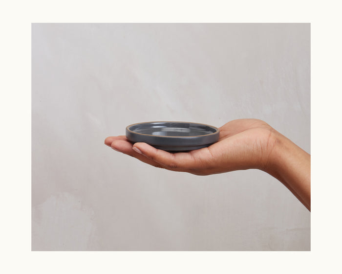A hand holds a Tiny Plates ceramic plate, flat and round, against a plain light background.