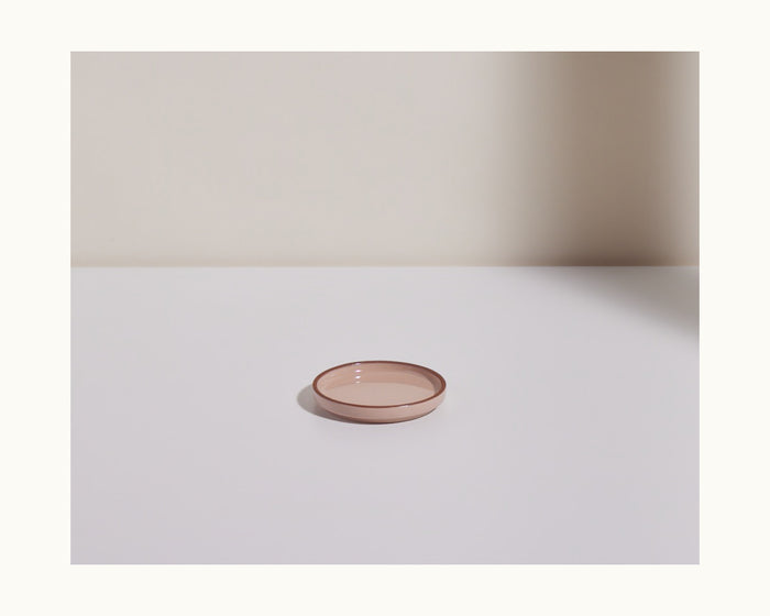 A Tiny Plates pink glass snack plate, small, round, and shallow, sits on a white surface with a beige wall and shadow in the background.