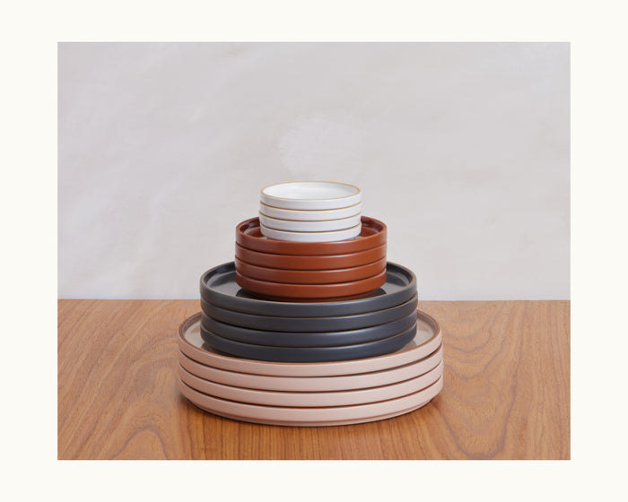 A stack of Tiny Plates, assorted ceramic pieces from a refined tableware set, arranged by size on a wooden surface with a light background.