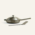 Stainless steel frying pan with lid and spatula on a white background