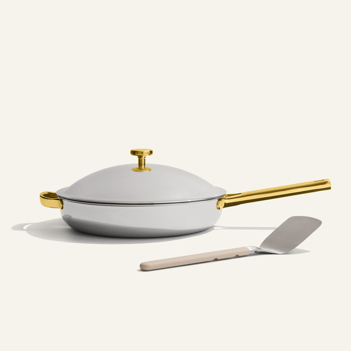The Titanium Always Pan Pro, a durable white sauté pan with lid and gold handle, sits behind a gray spatula with beige handle on a plain light background.